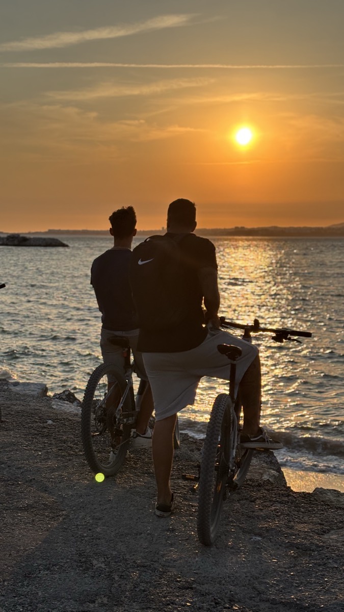 Two riders pause on a seafront breakwater to watch the sunset over the ocean at Costa da Caparica