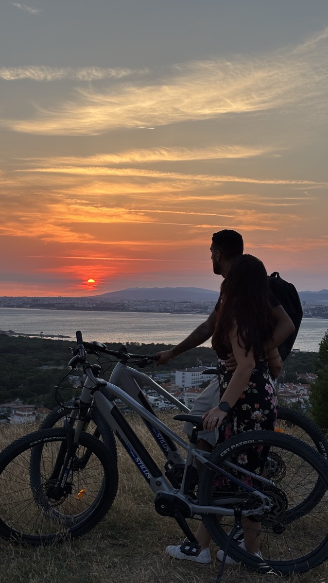 A couple with an eBike on a grassy hilltop overlooking the bay and Costa da Caparica at sunset