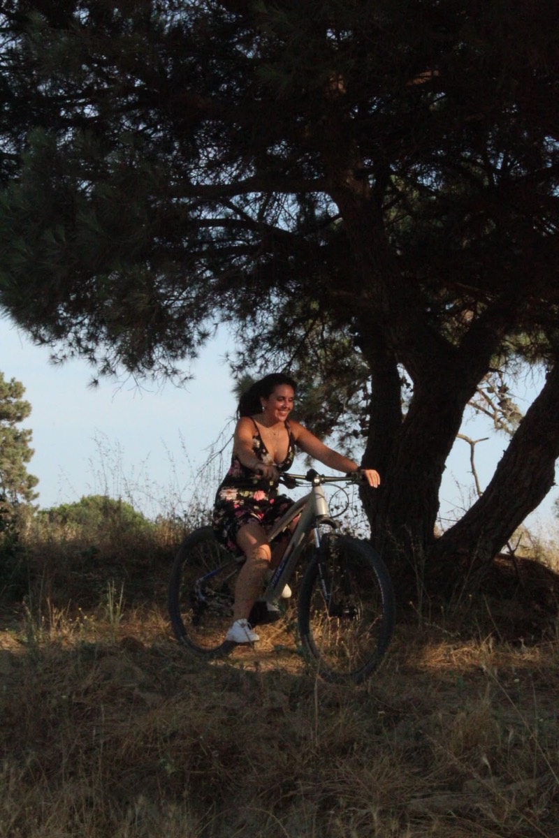 A smiling rider on an eBike under a large pine tree at golden hour near Costa da Caparica