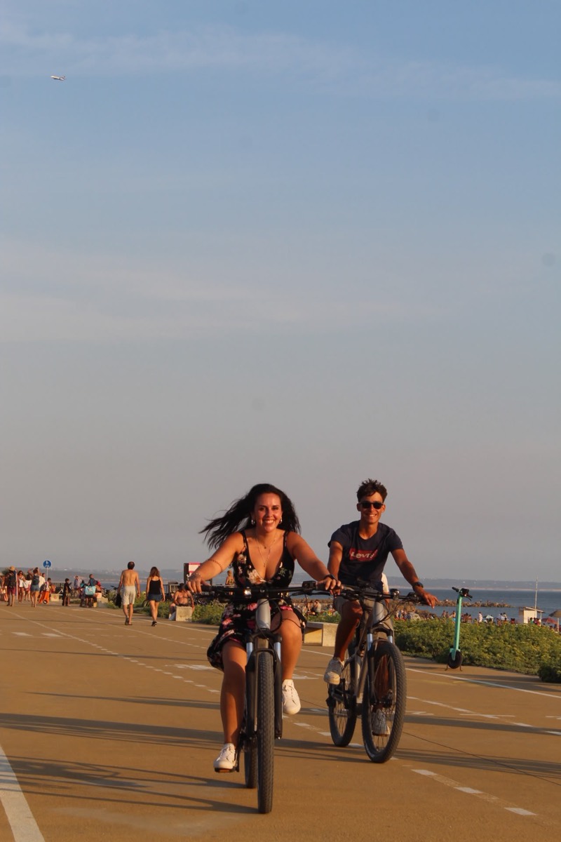 A smiling couple riding eBikes along the Costa da Caparica seafront promenade beside the beach
