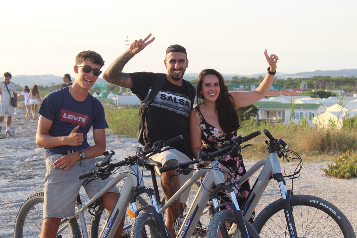 Three happy riders with thumbs up beside their eBikes near the Costa da Caparica beach