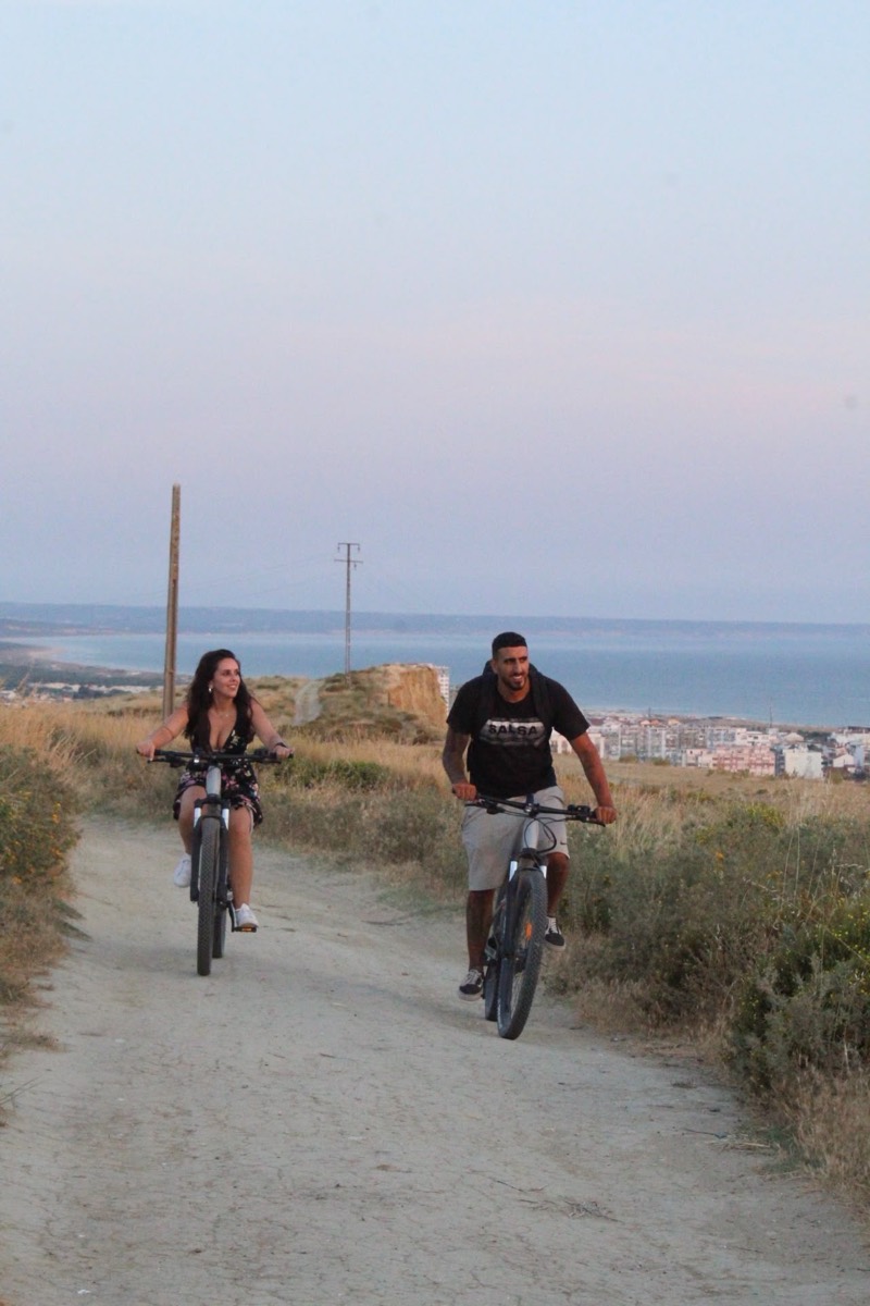 A couple riding eBikes up a coastal path with the town and coastline of Costa da Caparica behind
