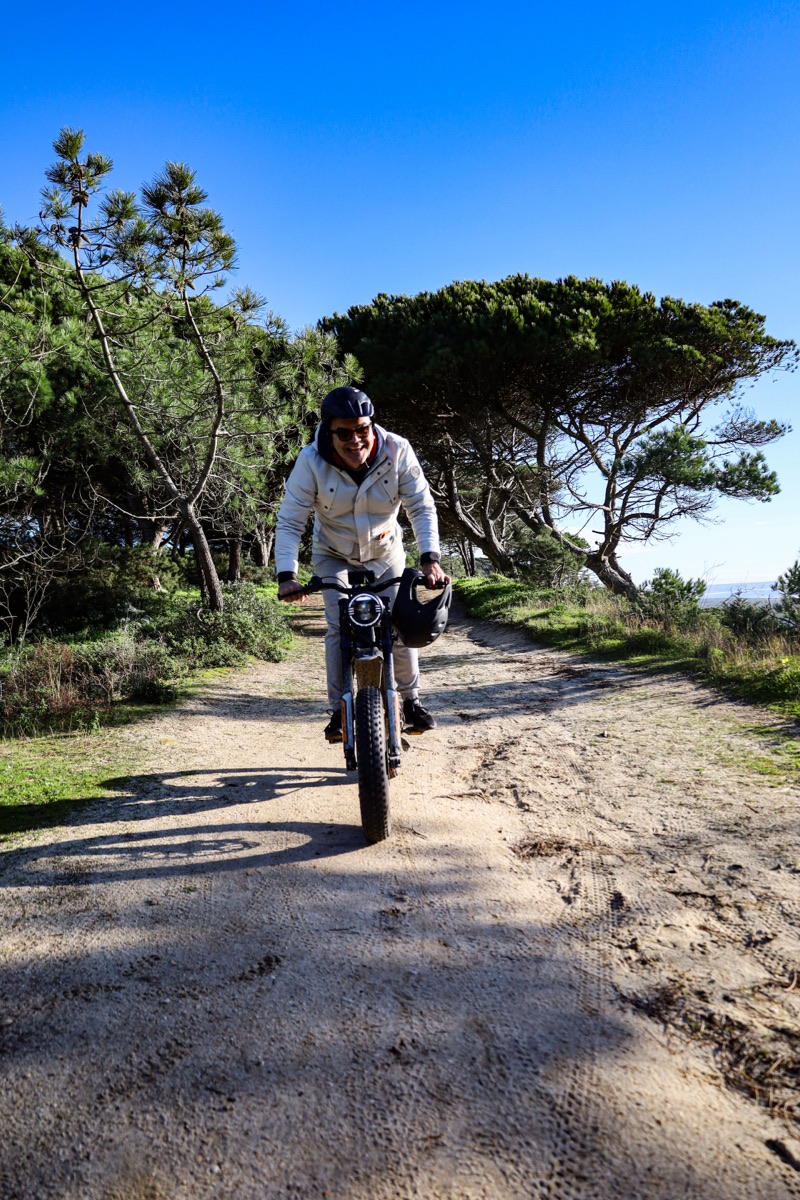 A rider heading toward the camera on an all-terrain eBike along a sunlit sandy pine-forest trail near Costa da Caparica