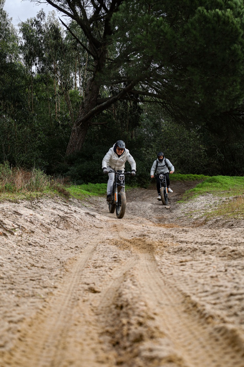 Two riders on all-terrain eBikes descending a sandy woodland track through pine and eucalyptus near Costa da Caparica
