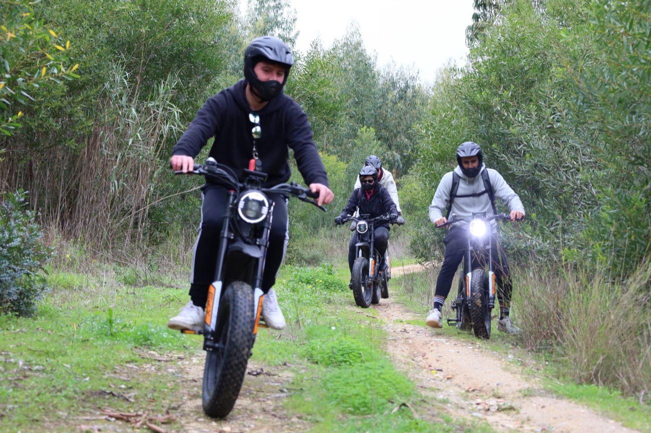 Three riders on all-terrain eBikes with headlights on along a narrow green woodland trail near Costa da Caparica