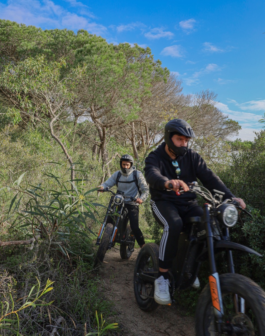 Two riders on all-terrain eBikes on a bushy forest trail under a blue sky near Costa da Caparica