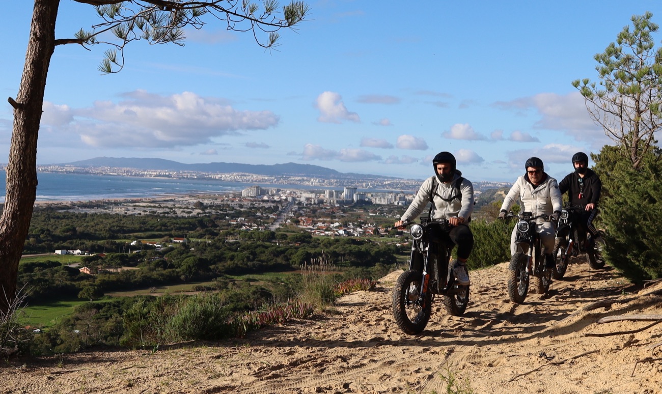 Three riders on eBikes descending a sandy hill with a panoramic view over the Costa da Caparica coastline