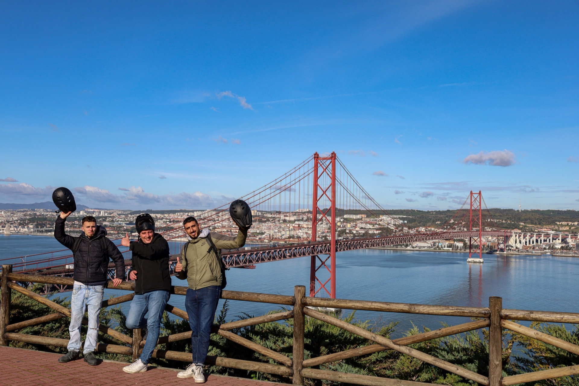 Three riders celebrating at an Almada viewpoint with the 25 de Abril Bridge and Lisbon behind