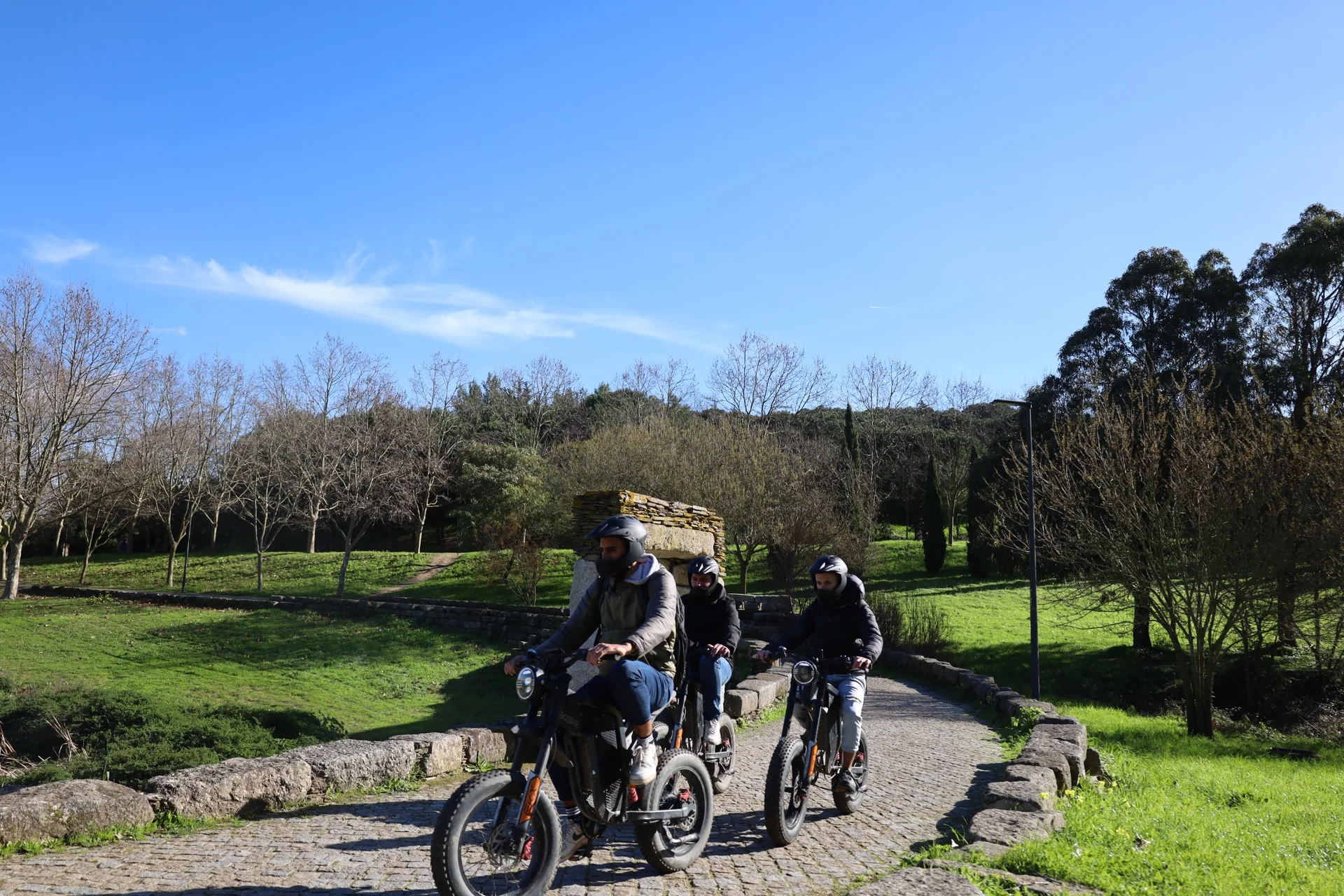 Three helmeted riders crossing a stone bridge through a green park in Almada