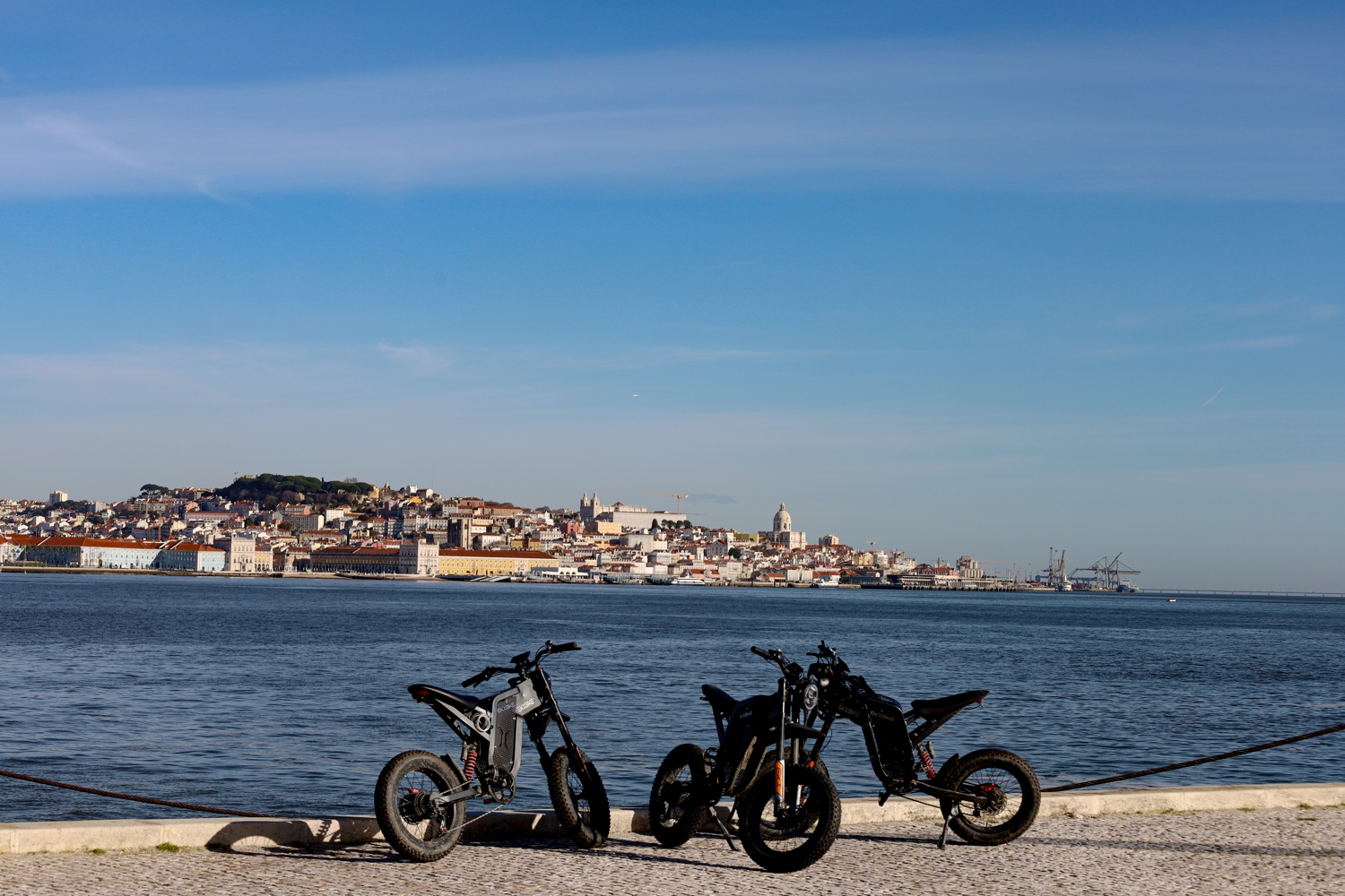 Two all-terrain eBikes parked on the Almada riverfront with the Lisbon skyline across the Tagus