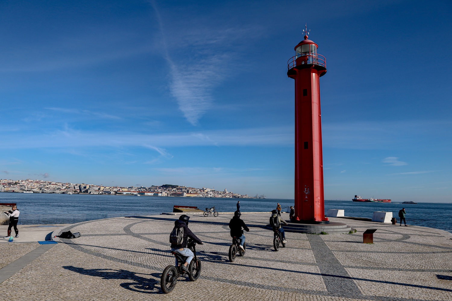 Riders cycling toward the red Cacilhas lighthouse in Almada with the Lisbon skyline across the river