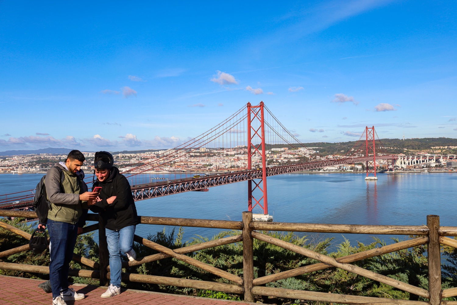 Two riders at an Almada viewpoint with the 25 de Abril Bridge and Lisbon behind