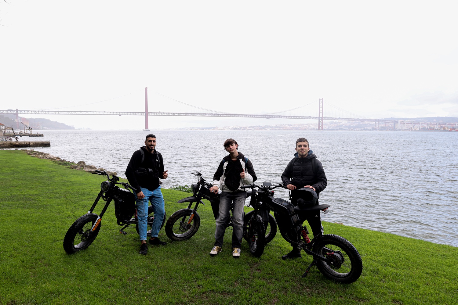 Three riders with their eBikes on the riverbank below the 25 de Abril Bridge in Almada